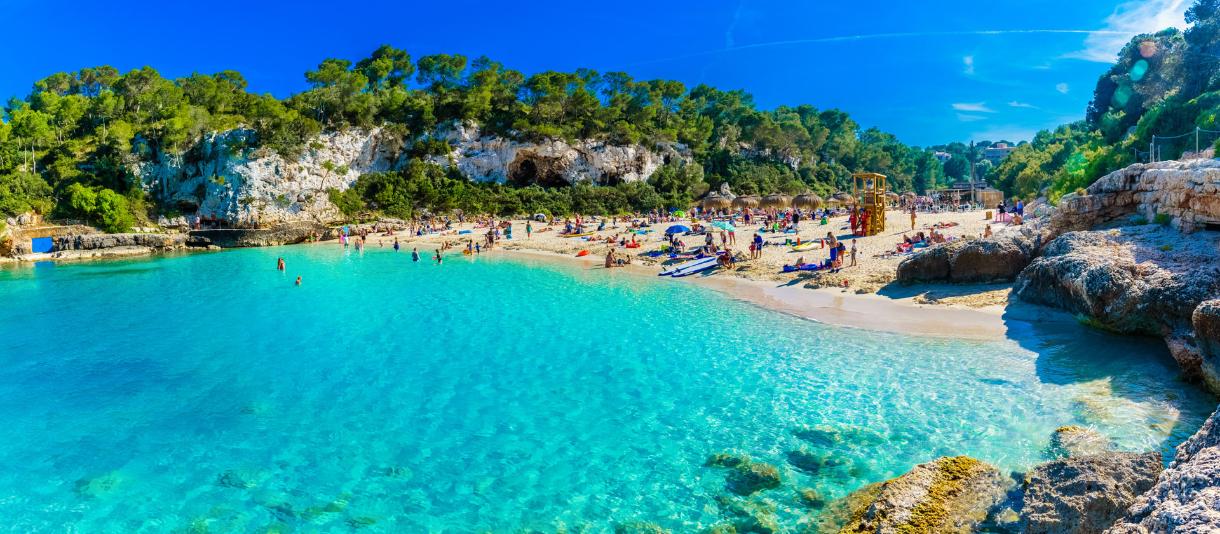 Blick auf Cala Llombards Strand auf Mallorca - türkisblaues Wasser mit Felsen und Bäumen auf beiden Seiten der Bucht