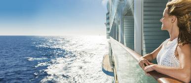 Frau steht an Geländer eines Kreuzfahrtschiffs mit Blick aufs offene Meer bei Sonnenschein.