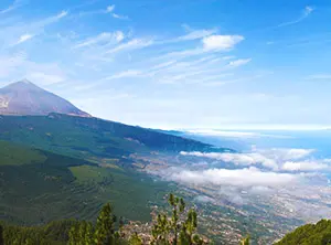Paysage volcanique de Tenerife avec le Teide, des pentes verdoyantes et un voile de nuages sous un ciel bleu clair.