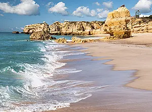 Plage de l’Algarve avec sable doré, falaises rocheuses ocre et vagues de l’océan Atlantique sous un ciel partiellement nuageux.