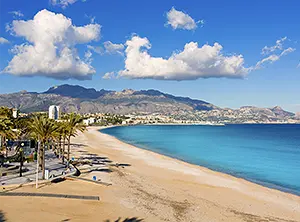 Plage de sable le long de la côte d’Alicante avec une promenade bordée de palmiers, la mer Méditerranée et des montagnes à l’arrière-plan.