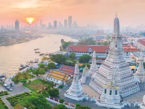 Vue sur le Wat Arun au bord du Chao Phraya à Bangkok, au lever du soleil, avec la skyline de la ville en arrière-plan.