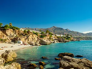 Felsige Badebucht an der Costa del Sol mit türkisfarbenem Meer, hellem Sandstrand und Bergen im Hintergrund.