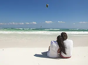 Paar entspannt am Strand, ein Kitesurfer gleitet über das Wasser.