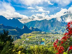 Blick auf das Dorf Cilaos im Talkessel Cirque de Cilaos auf La Réunion mit steilen Bergen, grünem Tal und roten Blumen im Vordergrund