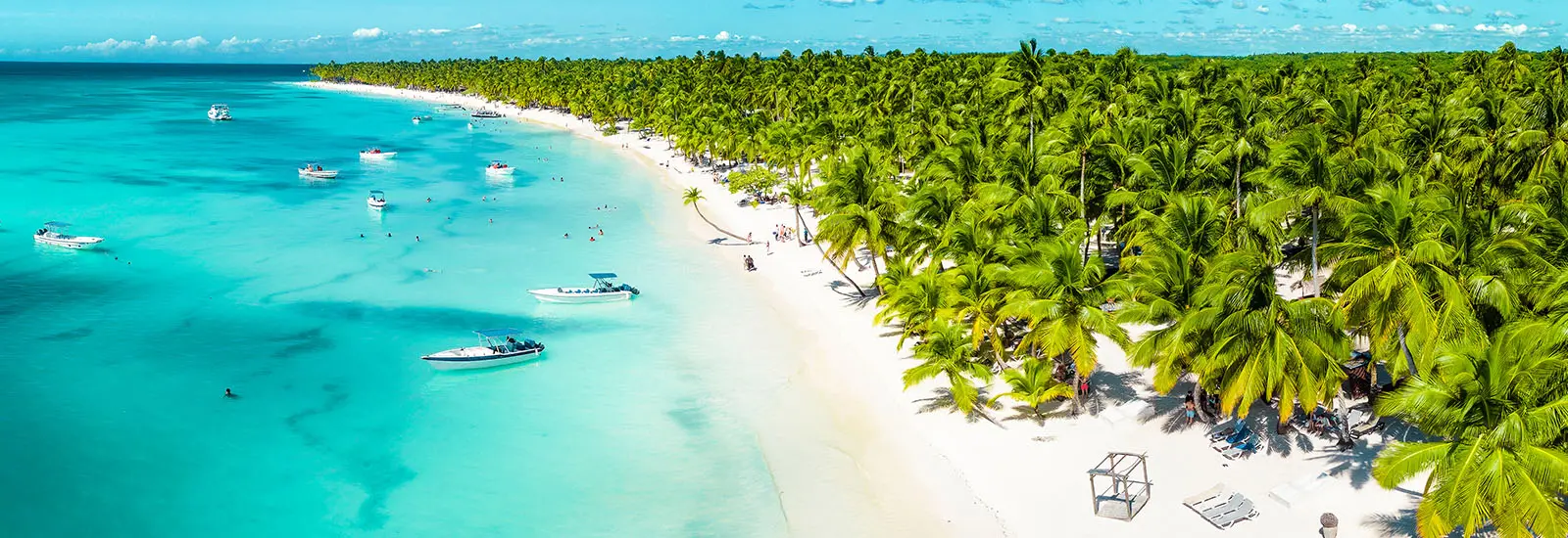 Vogelperspektive auf die Isla Saona: Weißer Sandstrand, dichte grüne Palmen und Boote im kristallklaren, türkisfarbenen Wasser.