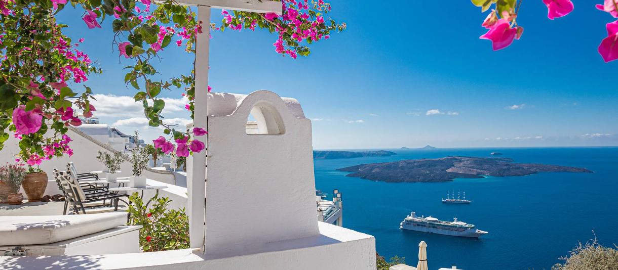Weiße Häuser mit pinken Bougainvillea und Blick auf die Caldera von Santorini mit Schiffen im Meer.