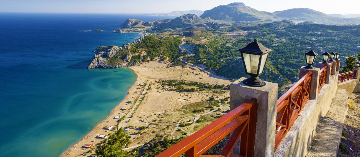 Rhodos Küstenpanorama mit türkisblauem Meer, Sandstrand, grüner Berglandschaft und Aussichtsterrasse im Vordergrund.
